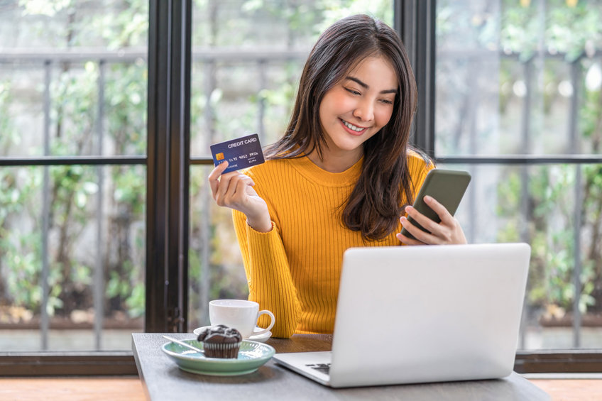 Young Asian woman using credit card and mobile phone for online shopping in coffee shop or coworking space, coffee cup, technology money wallet and online payment concept, credit card mockup